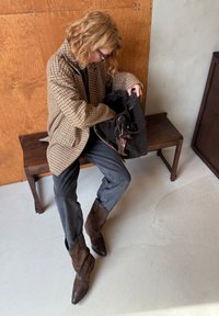 Blazer oversize a quadri marroni, camicia nera, jeans grigi e stivaletti in suede marroni con dettagli a stella, seduto su una panchina di legno.
