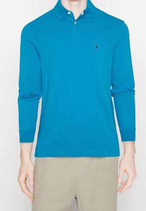 Mann trägt ein leuchtend blaues langärmeliges Polohemd und beige Hose, steht vor einem schlichten hellen Hintergrund.