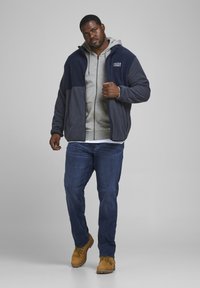 Veste en polaire bleu marine et gris avec une capuche sur un sweat à capuche gris clair, associée à un jean bleu et des bottes de travail beiges. Position frontale, attitude décontractée.