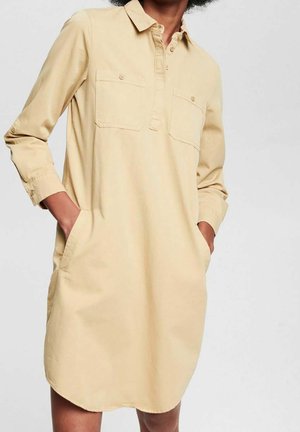 Beige long-sleeve shirt dress with front pockets, collar, and buttoned placket worn with hands in side pockets.