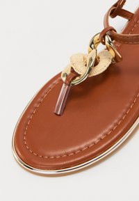 Sandalia de cuero marrón con acento de cadena dorada, detalle tejido beige y una correa transparente. Presenta una suela suave y redondeada con ribete metálico.