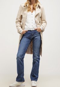 Beige trench coat, jossa on kaksirivinen malli, käytetty valkoisen teksturoidun yläosan ja sinisten farkkujen päällä. Viimeistely valkoisilla lenkkareilla.