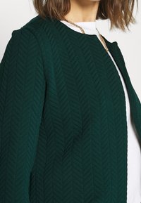 Tummanvihreä tikattu takki, jossa on herringbone-kuvio, pyöreä kaula-aukko ja teksturoitu kangas. Päälle puettuna valkoinen paita.
