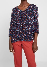Blommig blus med marinblå bakgrund, som har röda, blå och vita detaljer. V-ringning och trekvartslånga ärmar. Lättviktsmaterial.