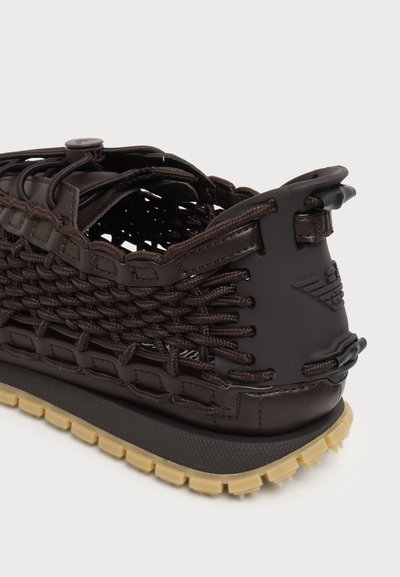 Sandale tissée marron foncé avec lacets réglables, semelle en caoutchouc et bande de roulement beige texturée mettant l’accent sur le talon et les détails latéraux.