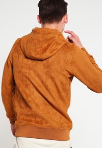 Sudadera con capucha marrón de textura suave y similar al ante. Cuenta con un bolsillo canguro y puños y dobladillo acanalados. La vista trasera muestra la capucha.