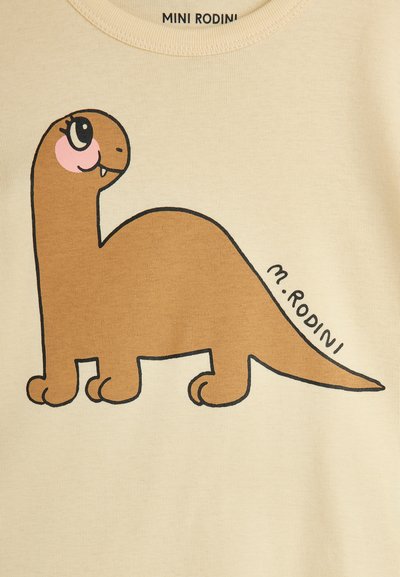 Beiges T-Shirt mit einem braunen Cartoon-Dinosaurier-Design, das große Augen und einen dezenten Schimmer hat. Eine schwarze Umrandung akzentuiert das Design.