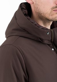 Giacca marrone con cappuccio, caratterizzata da una texture esterna liscia, con zip e chiusure a scatto. L'interno del cappuccio presenta un colore a contrasto.