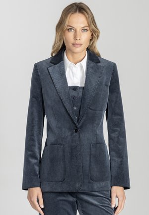 Kobieta ubrana w niebieski garnitur z korduroju z marynarką zapinaną na jeden guzik, dopasowanymi spodniami i białą koszulą z kołnierzykiem, stojąca na jednolitym tle.