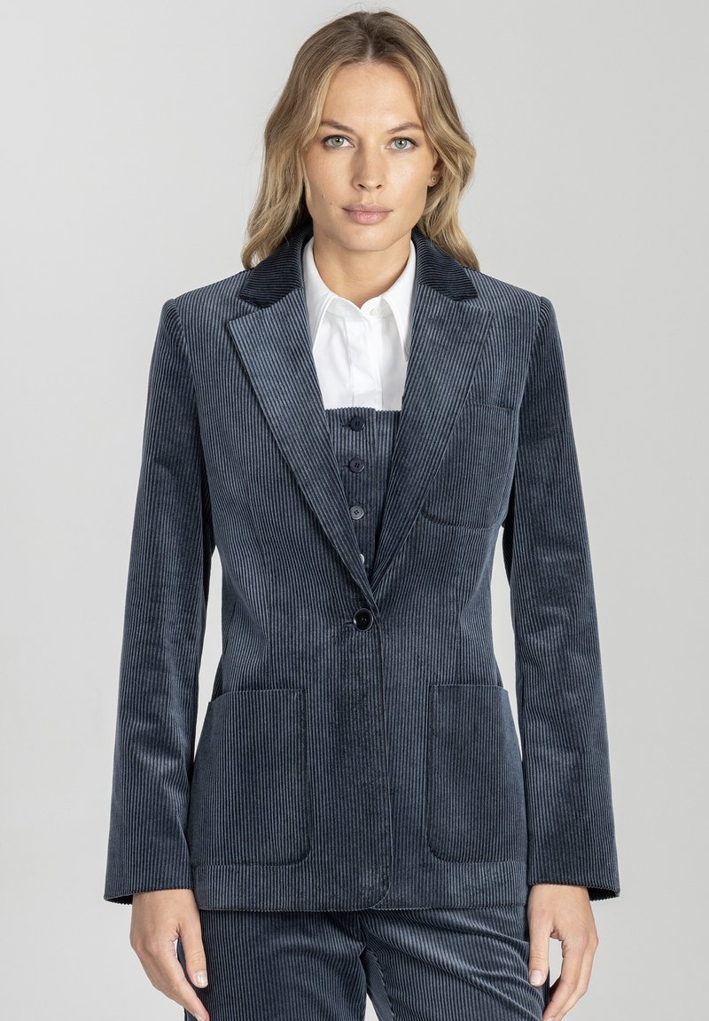 Kobieta ubrana w niebieski garnitur z korduroju z marynarką zapinaną na jeden guzik, dopasowanymi spodniami i białą koszulą z kołnierzykiem, stojąca na jednolitym tle.