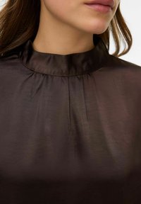 Blusa marrón oscuro con cuello alto y pliegues sutiles. La tela tiene una textura suave y brillante, que crea una apariencia elegante.