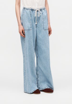 Person iført løstsiddende lyseblå denim bukser med store forlommer og en hvid snøre, kombineret med brune sko.