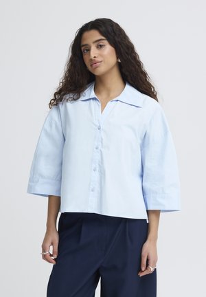 Žena s dlhými kučeravými vlasmi, oblečená v svetlomodrej veľkej košeli s golierom a tmavomodrých nohaviciach, stojaca na jednotnom pozadí.