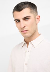 Jasnobeżowa lniana koszula z kołnierzykiem zapinanym na guziki, z małymi kremowymi guzikami i subtelnymi haftami na klatce piersiowej.