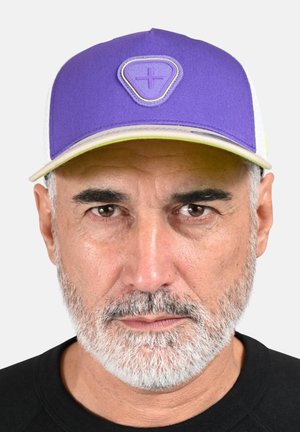 Homme d'âge moyen avec une barbe grise portant une casquette violette et blanche avec un emblème triangulaire affichant un signe plus, expression neutre, chemise noire.