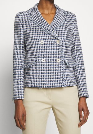 Femme portant un blazer croisé à motif pied-de-poule bleu et blanc avec des détails en franges et un pantalon beige.