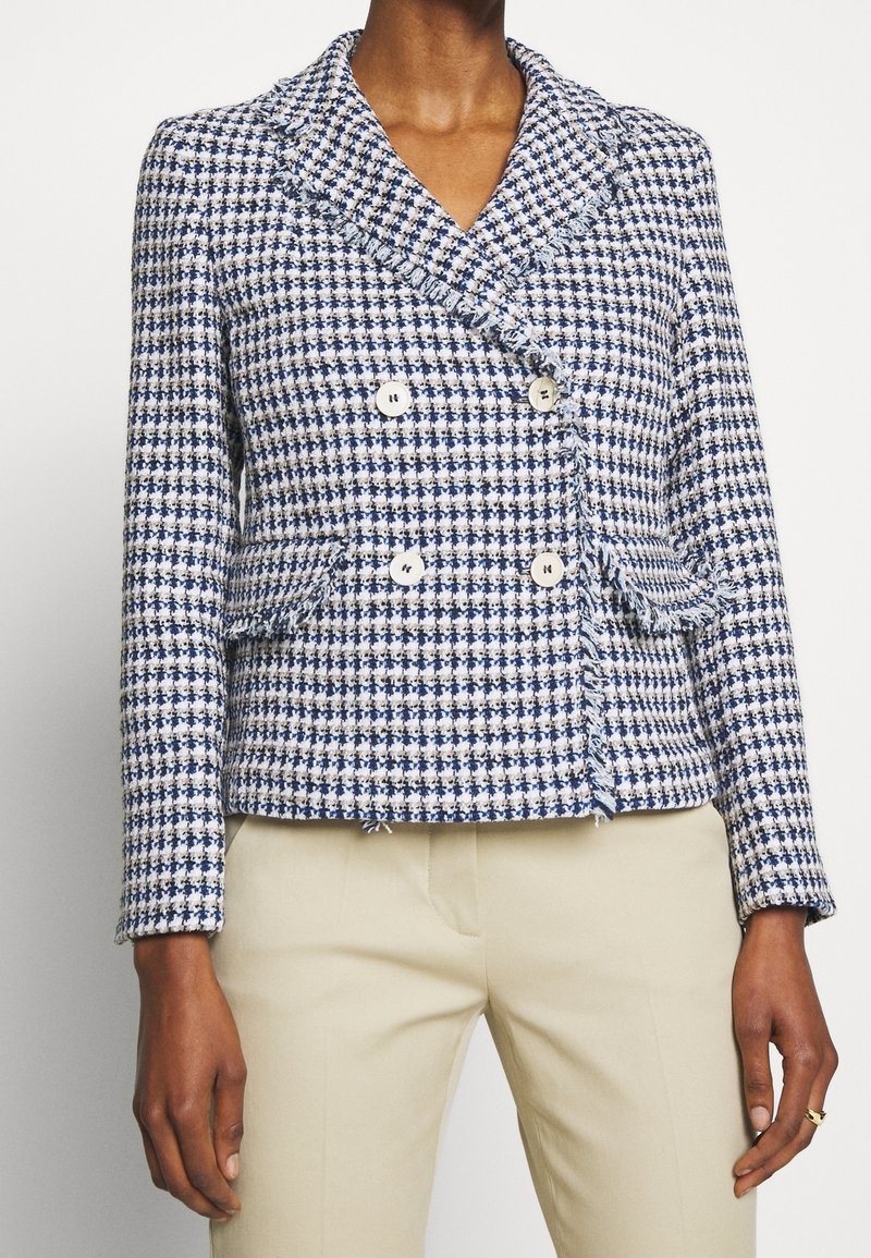 Femme portant un blazer croisé à motif pied-de-poule bleu et blanc avec des détails en franges et un pantalon beige.