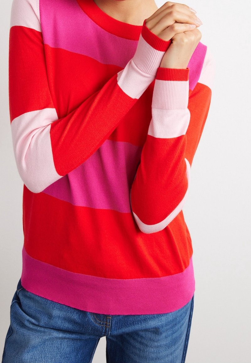 Personne portant un pull rayé avec des bandes rouges, roses et rose clair, tenant un poignet avec les deux mains, jean bleu visible.