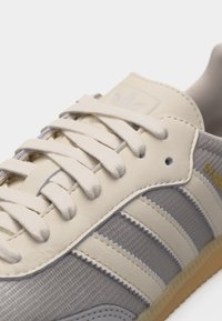 Κοντινή λήψη από αθλητικό παπούτσι Adidas σε μπεζ και γκρι αποχρώσεις με λευκά κορδόνια, τρεις λευκές ρίγες και χρυσή επιγραφή "SAMOA" στο πλάι.