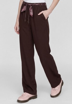 Person wearing loose dark brown pants with a patterned fabric belt, one hand in pocket, and pink tassel loafers on a light background.