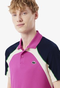 Pikétröja i rosa med mörkblå ärmar och ljusbeige meshdetaljer, med gröna accenter och en liten broderad logotyp på bröstet.