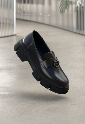 Mocasín de cuero negro con suela con textura gruesa y hebilla plateada, exhibido sobre un suelo de hormigón liso y un fondo de barras de metal verticales.