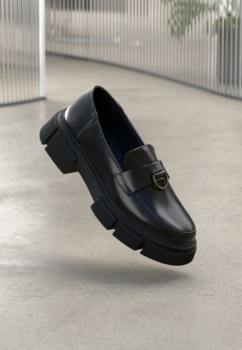 Mocassin en cuir noir avec une semelle épaisse texturée et une boucle argentée, présenté sur un sol en béton lisse et un fond de barreaux métalliques verticaux.