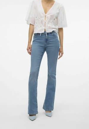 Lichte spijkerbroeken met flair en een aansluitende taille, gecombineerd met een witte, korte blouse met oogjes en een strik aan de voorkant. Gecombineerd met blauwe hakken.