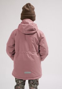 Roze waterdichte jas met een capuchon, verstelbare taille en een gladde textuur, gecombineerd met groenachtige geblokte broek en een bruine gebreide beanie.