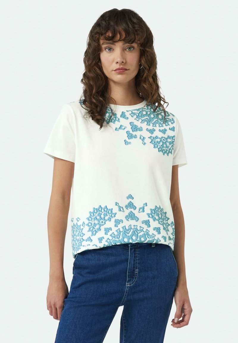 Witte katoenen T-shirt met ingewikkelde lichtblauwe bloemenborduring op de borst en onderrand, met een ronde halslijn en korte mouwen.