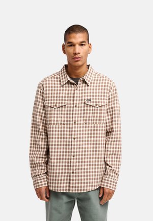 Jeune homme portant une chemise à carreaux marron et blanche avec deux poches poitrine, debout sur un fond clair uni.