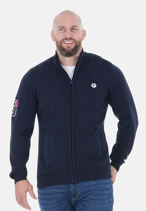 Cardigan zippé marine en tissu texturé, avec poches latérales et un patch logo sur la poitrine gauche. Coupe classique avec des poignets côtelés.