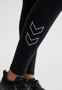 Schwarze sportliche Leggings mit glatter Textur, versehen mit einem weißen Chevron-Muster an der Seite und kombiniert mit schwarzen Sneakers.