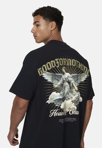 T-shirt in cotone nero con una grande grafica di un angelo sul retro, con testo dorato "GOODFORNOTHING" e raggi che si irradiano da esso.