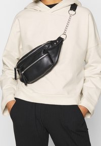 Riñonera de cuero negro con dos compartimentos con cremallera, correa de cadena y tela, combinada con una sudadera ligera con capucha y pantalones negros con costillas.