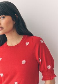 Woman with long dark hair wearing a red short-sleeve sweater with white daisy patterns and smocked cuff detail.