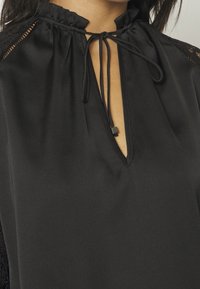 Blouse en satin noir avec un col montant fermé par un lien, des accents en dentelle sur les épaules et une ouverture en forme de goutte d'eau à l'avant.