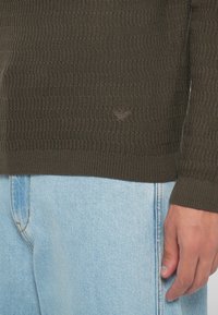 Tume oliivroheline kudum, millel on soonikkoe tekstuur ja väike kolmnurkne logo detail. Koos helesiniste denim-pükstega.