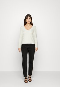 Lichtbeige cardigan met een V-hals en lange mouwen, voorzien van een knoopsluiting aan de voorkant, gecombineerd met zwarte rechte jeans en platte sandalen.