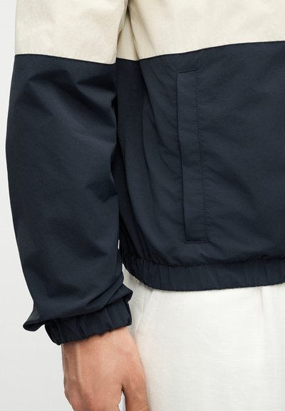 Κοντινό πλάνο ενός ατόμου που φοράει ένα μαύρο και μπεζ μπουφάν με ελαστική μανσέτα και πλαϊνή τσέπη, σε συνδυασμό με ανοιχτόχρωμο παντελόνι.