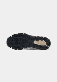 Running shoe outsole in black rubber with a patterned tread design and beige accents, showcasing textured grooves and a smooth central area.