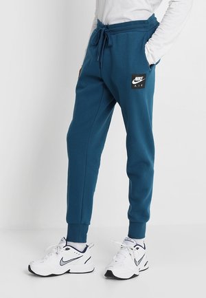 Personne portant un jogging Nike Air bleu canard et des baskets blanches Nike Monarch, debout les mains dans les poches.