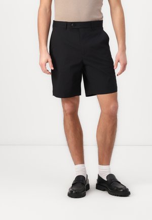 Homme portant un short noir mi-genou, des chaussettes blanches, des mocassins noirs et un haut beige sans manches, debout devant un fond blanc uni.