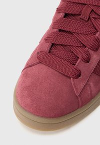 Bordo spalvos zomšiniai sportbačiai su tekstūruotais batraiščiais, apvalia nosyte ir guminiais padais. Turi siuvinėtus detalių ir sportišką dizainą.