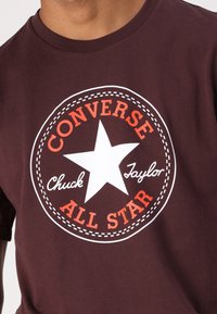 T-shirt en coton bordeaux avec un graphique circulaire blanc et orange comportant une étoile et le texte "Converse Chuck Taylor All Star".