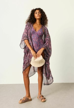 Woman wearing a purple paisley swimsuit and matching cover-up, holding a beige woven hat, standing barefoot on a light floor against a plain wall.