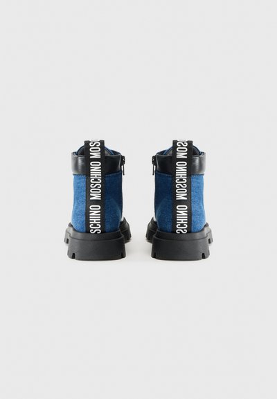 Botas tobilleras de tela azul con acentos de cuero negro, suela negra gruesa y una tira de logotipo blanca prominente en la parte posterior. Detalle de cremallera en los laterales.