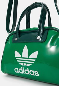 Zelená lakovaná kabelka zaobleného tvaru, s dvojitými uchy a odnímatelným popruhem. Na přední straně se nachází bílé logo Adidas se třemi pruhy.