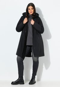 Jeune femme portant un manteau d'hiver noir à capuche, un col roulé sombre, un jean noir et des bottes noires, se tenant devant un fond blanc.