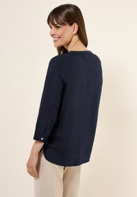 Blusa blu navy a maniche ¾ in tessuto leggero con scollo rotondo e orlo curvo, dotata di polsini con bottoni e una superficie liscia.